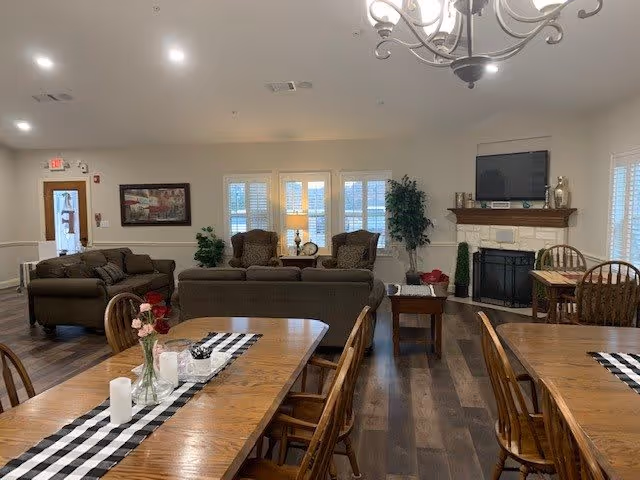 A cozy common area in Mustang Creek Estates Frisco featuring wooden dining tables with checkered runners and flower vases, comfortable sofas and armchairs arranged around a coffee table, a fireplace with a TV mounted above it, and large windows with white shutters letting in natural light.