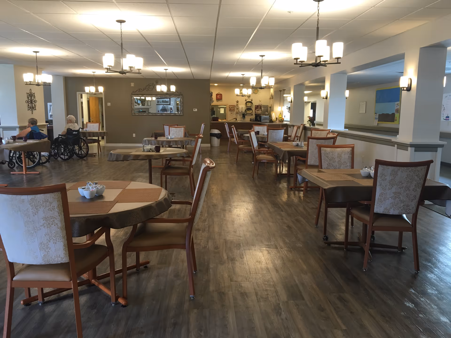 Spacious assisted-living dining room with multiple tables and chairs, chandeliers, wood flooring, and two residents in wheelchairs at the back left.