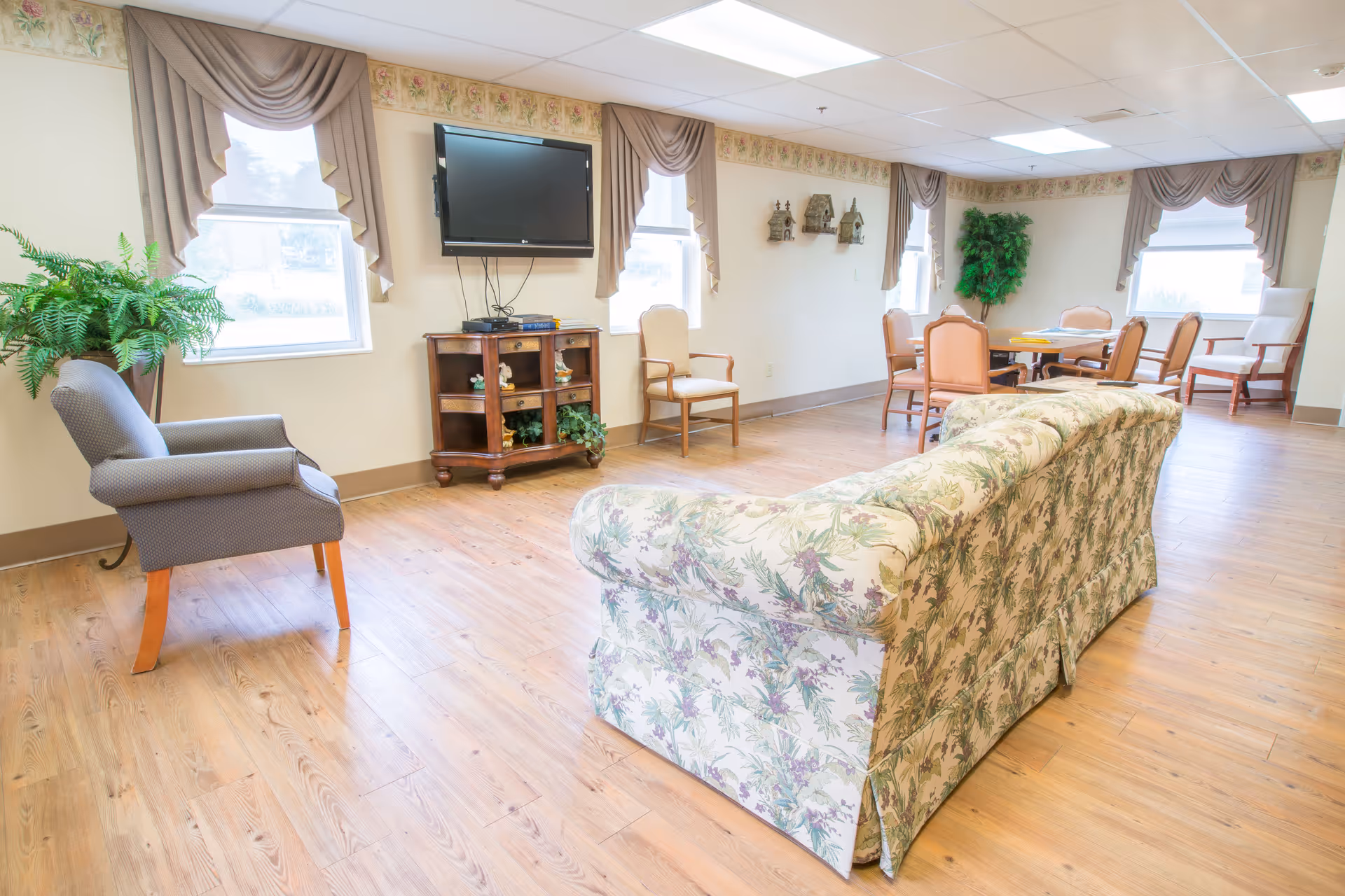 A bright and spacious common area in a senior living facility with wooden flooring, floral-patterned sofa, several chairs, a wall-mounted TV, and decorative plants near the windows.