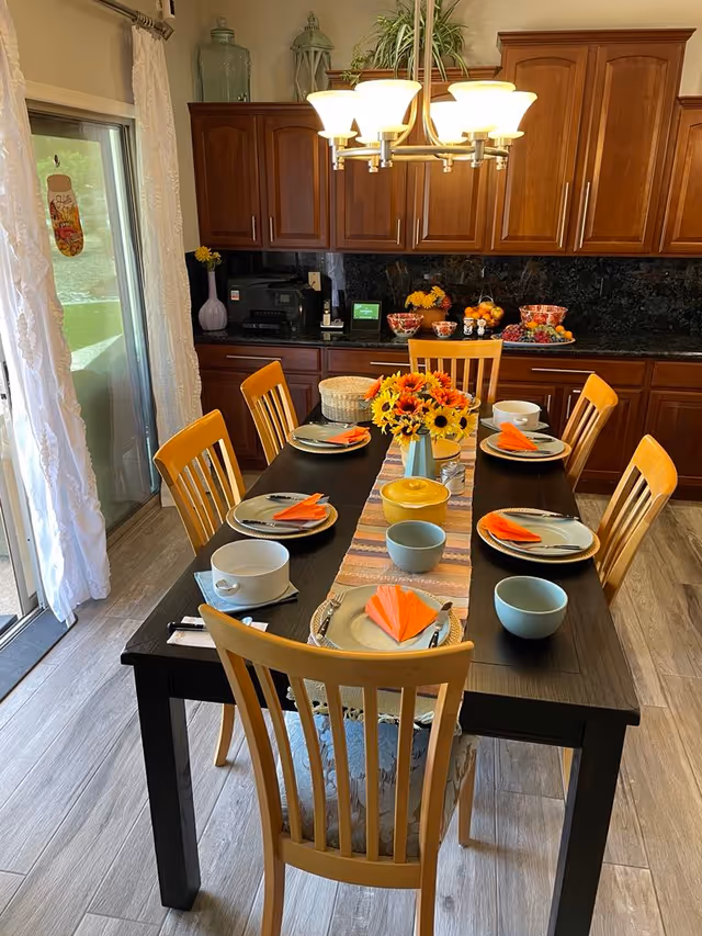 A dining room with a dark wooden table set for six people. Each place setting has a plate, bowl, utensils, and an orange folded napkin. A colorful table runner and a vase with sunflowers decorate the center of the table. Behind the table are wooden cabinets and a countertop with bowls of fruit and other items. A chandelier with six lights hangs above the table. To the left, there is a sliding glass door with white curtains.