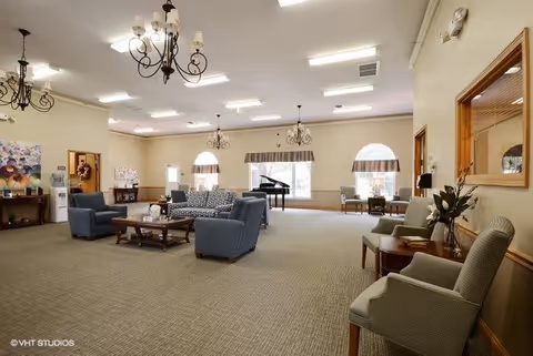 A spacious and well-lit living room area with multiple seating arrangements including blue armchairs and a patterned sofa around a wooden coffee table. The room features chandeliers hanging from the ceiling, large windows with arched tops and valances, a grand piano near the windows, and additional chairs along the walls. The walls are painted light beige with wooden trim, and there are decorative elements such as flowers and artwork.