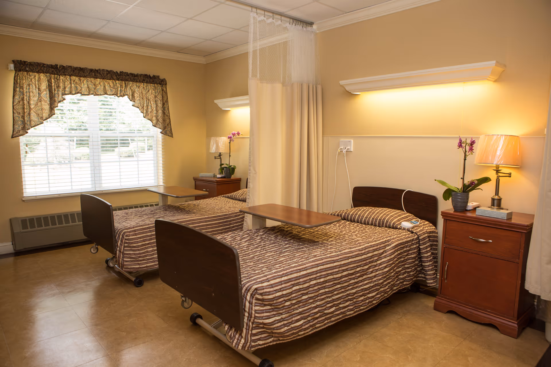A room with two hospital-style beds separated by a privacy curtain. Each bed has a brown striped bedspread and an overbed table. There are two wooden nightstands, each with a lamp and a potted orchid. A large window with blinds and a decorative valance lets in natural light. The walls are painted beige and the floor has a light brown tile.