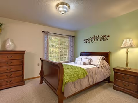 A cozy bedroom with a wooden sleigh bed dressed in beige bedding and a green throw blanket. There is a wooden dresser on the left and a matching nightstand with a lamp on the right. A window with curtains lets in natural light, and a decorative wall piece hangs above the bed.