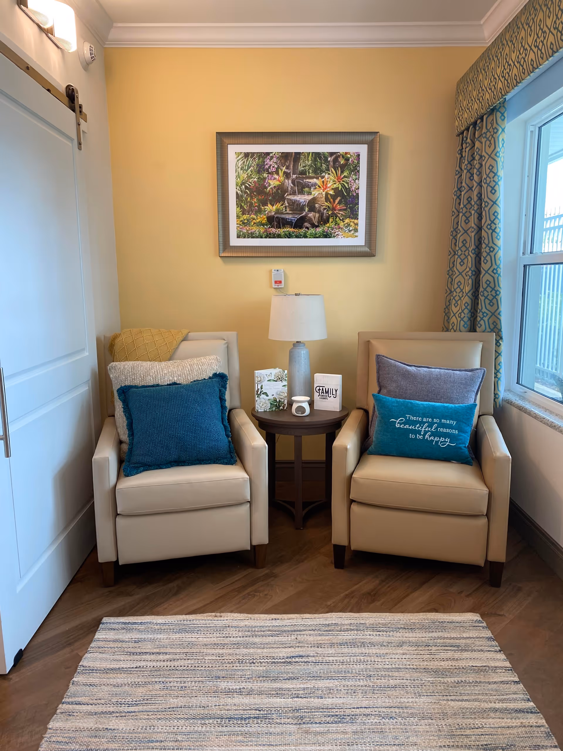 A cozy seating area with two beige armchairs, each adorned with multiple decorative pillows in shades of blue, gray, and yellow. Between the chairs is a small round wooden table with a lamp and two greeting cards. Above the table hangs a framed picture of a waterfall surrounded by colorful flowers. To the right is a window with patterned blue and beige curtains, and to the left is a white sliding barn door. The floor is wooden with a light-colored woven rug in front of the chairs.