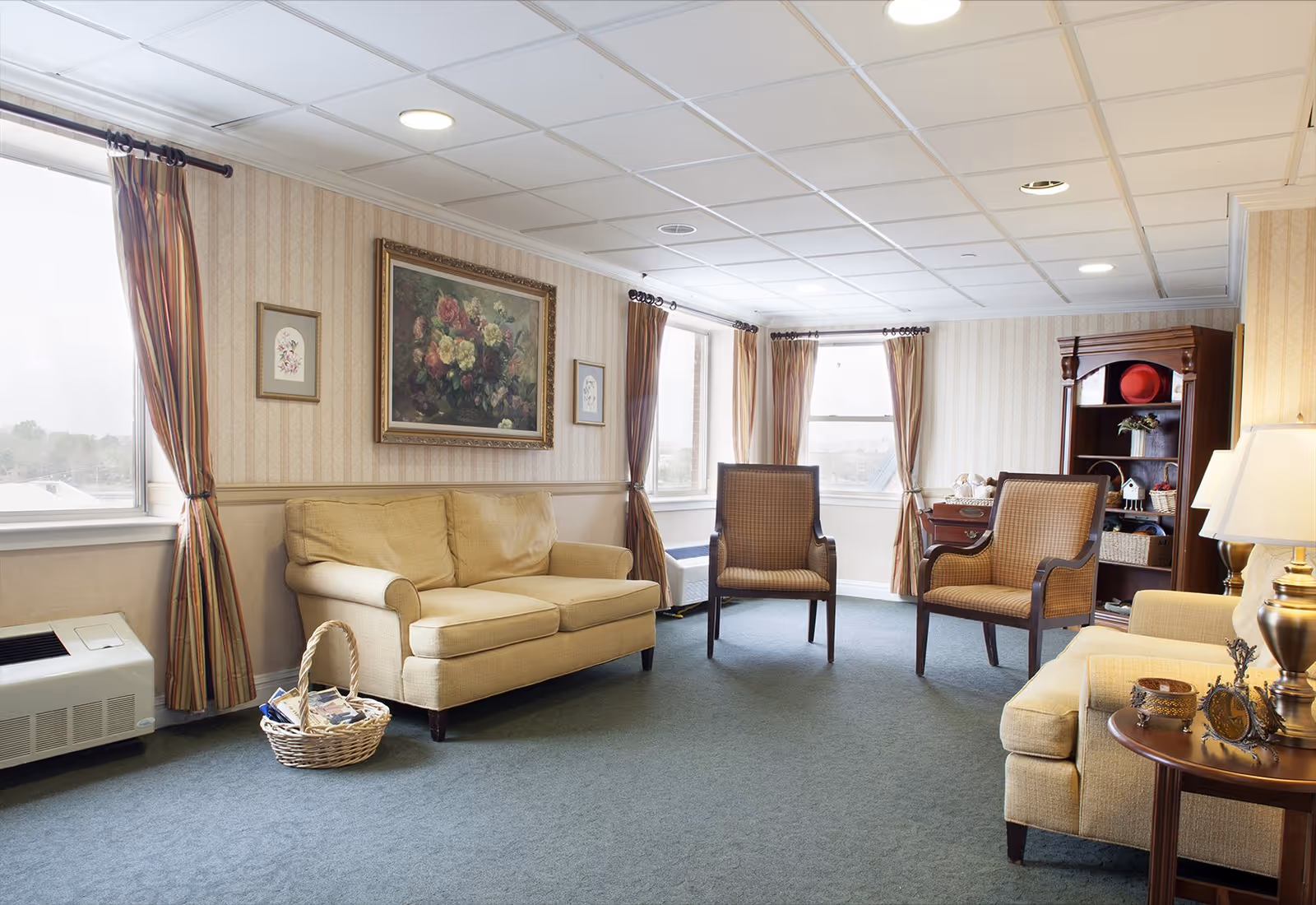 A cozy living room with beige sofas and two wooden armchairs with patterned cushions. The room has three windows with striped curtains, a blue carpet, and wallpaper with a subtle pattern. A large floral painting and two smaller framed pictures hang on the wall. There is a wooden bookshelf with decorative items and a side table with a lamp and small decorative objects.