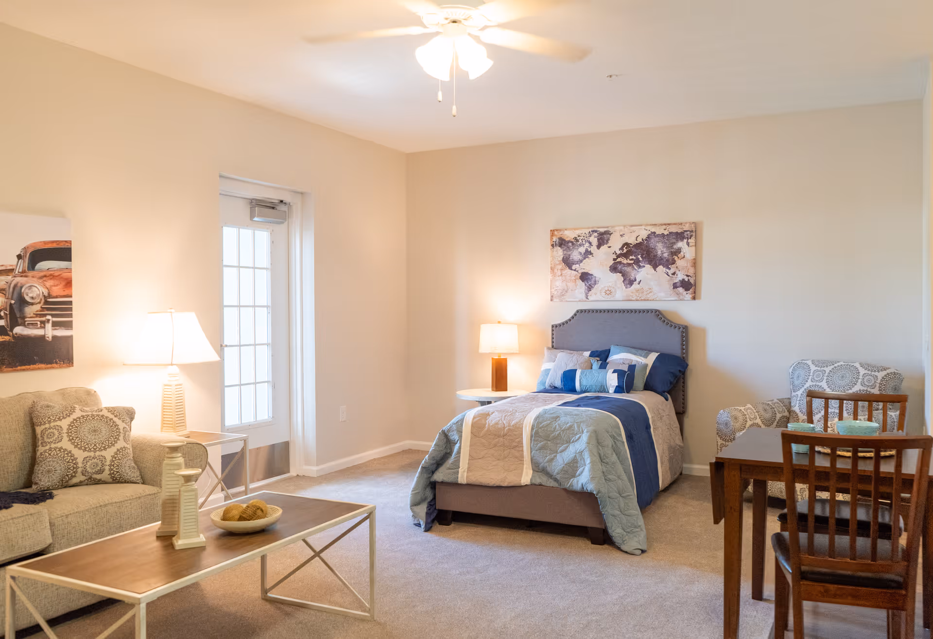 A cozy senior living room featuring a bed with blue and beige bedding, a gray upholstered headboard, a wooden dining table with chairs, a patterned armchair, a beige sofa with decorative pillows, a coffee table with decorative items, two table lamps, a ceiling fan, and a world map artwork on the wall.