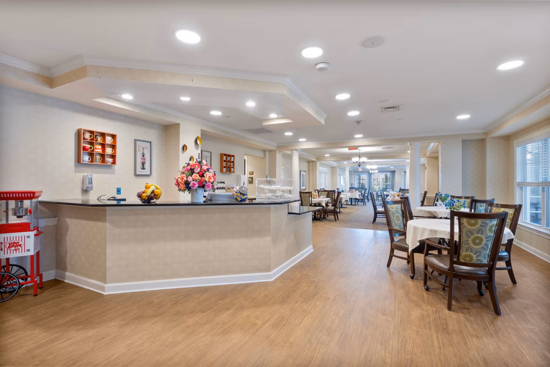 A spacious dining area in a senior living facility with multiple round tables covered with white tablecloths and wooden chairs with patterned cushions. There is a counter with a flower vase, a fruit bowl, and a popcorn machine on the left side. The room has wood flooring, recessed ceiling lights, and large windows letting in natural light.