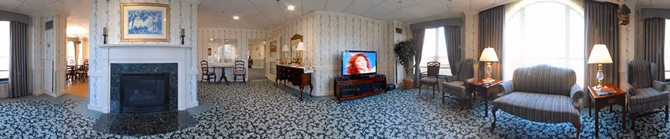 Panoramic view of a senior living facility common area featuring a fireplace with a framed painting above it, patterned carpet, wallpapered walls, a television on a wooden stand, several chairs and small tables with lamps, and large windows with curtains allowing natural light.