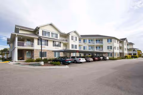 Exterior view of a three-story retirement community building with balconies and a covered parking area in front. Several cars are parked in the parking lot, and the sky is partly cloudy.