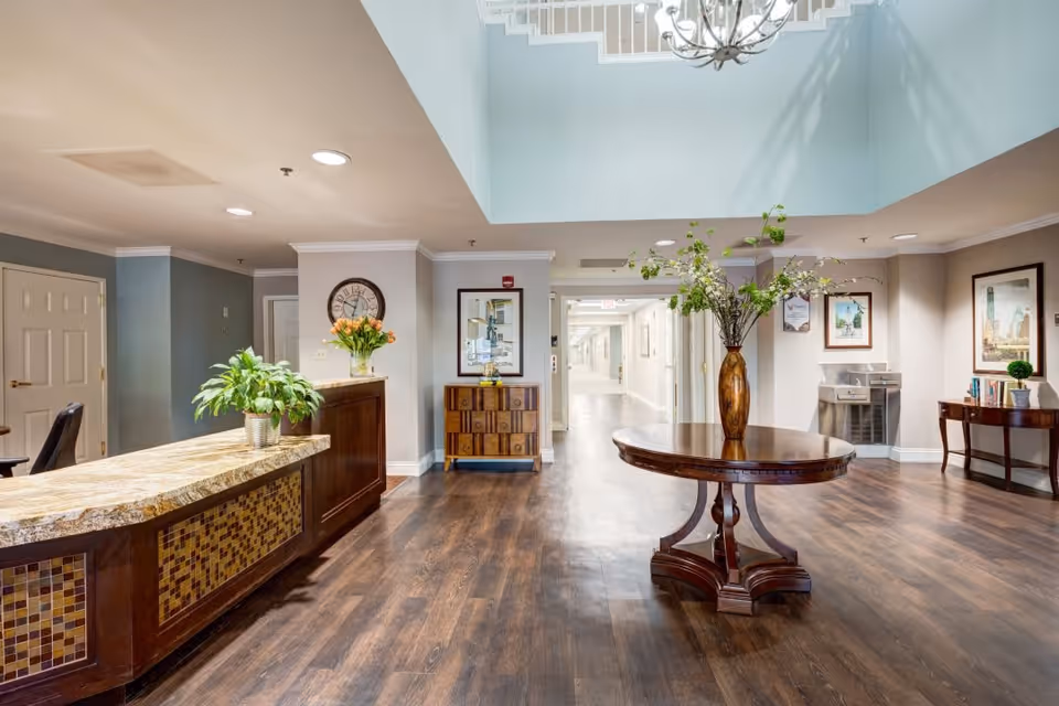 A spacious and well-lit interior lobby area with a wooden reception desk on the left, a round wooden table with a large vase of flowers in the center, and framed artwork on the walls. The floor is dark wood, and there is a hallway leading further into the building.
