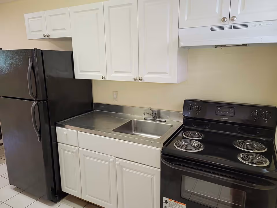 A kitchen area with white cabinets, a stainless steel sink, a black electric stove with four coil burners, and a black refrigerator. The countertop is stainless steel and the walls are painted light beige.