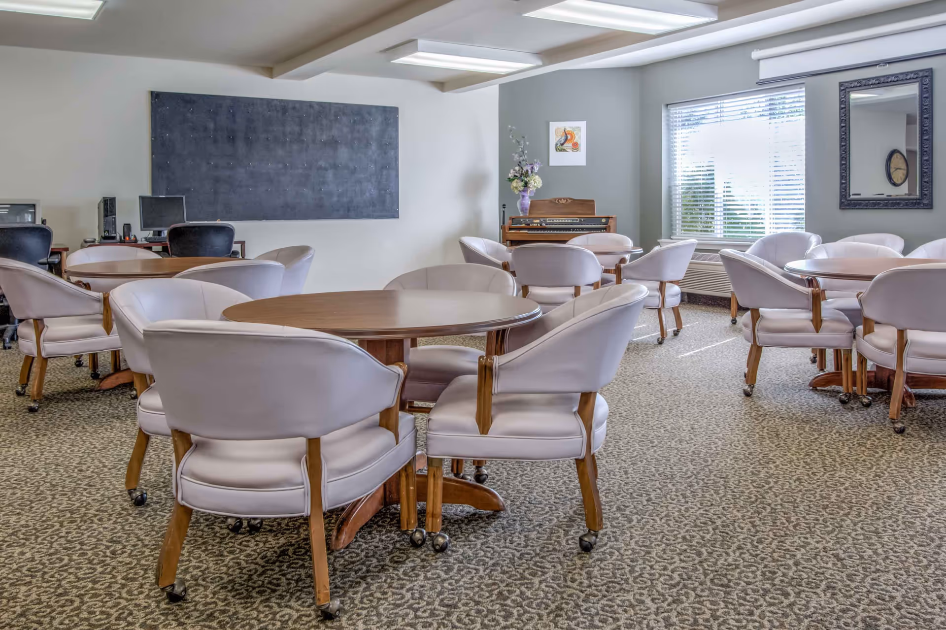 Bright common room with multiple round tables and upholstered chairs on patterned carpet, a piano and windows along one wall.