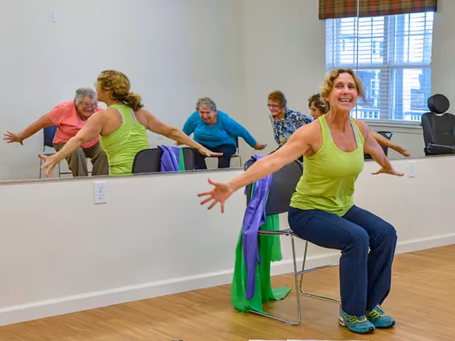 A group of elderly women participating in a seated exercise class in a bright room with wooden floors and a large mirror on the wall. One woman in a green tank top and blue pants is seated on a chair with arms extended, smiling. Other women are visible in the mirror, also engaged in the exercise.