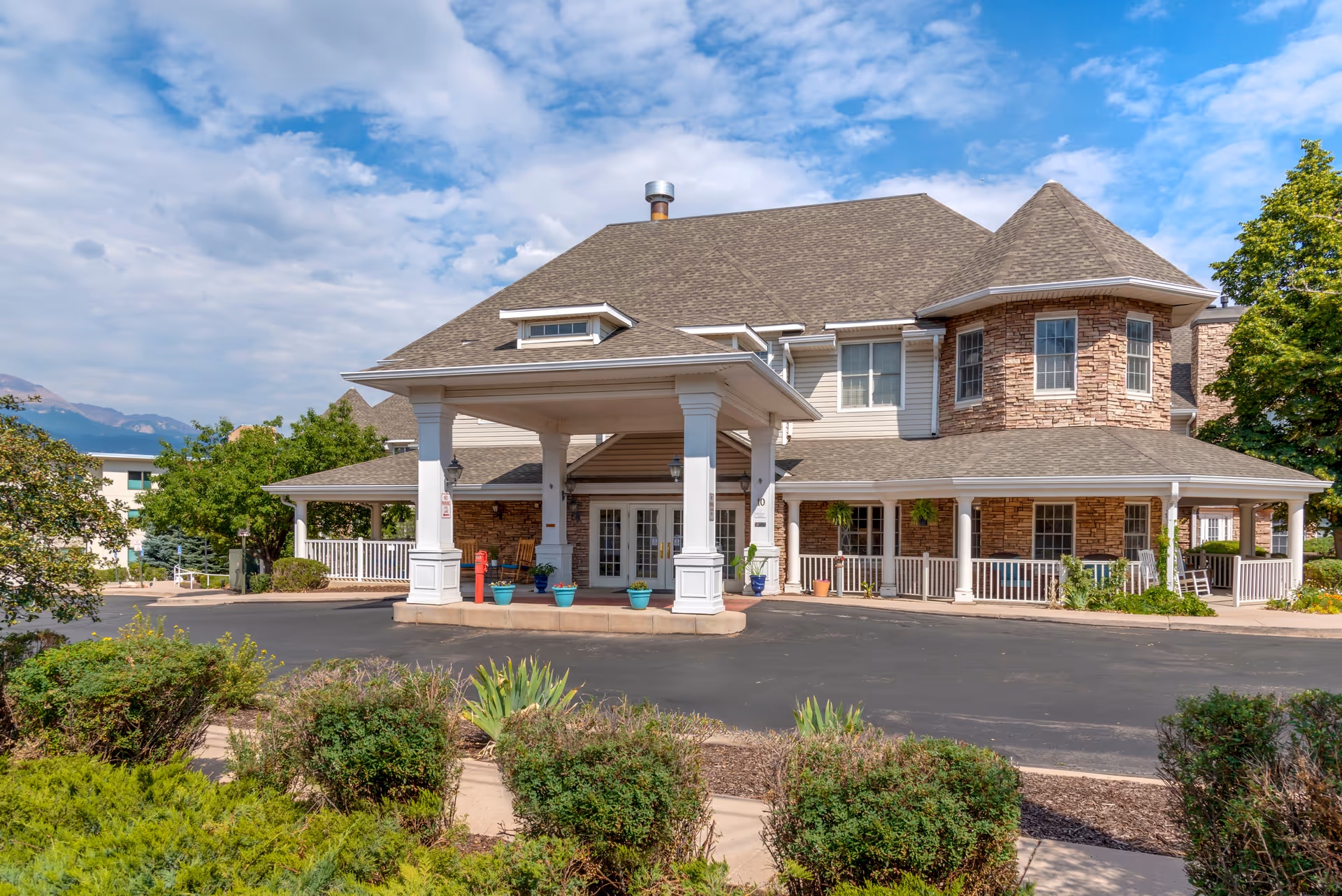 Exterior view of a senior living facility building with a covered entrance, stone and siding facade, multiple windows, and a wrap-around porch with rocking chairs. There are shrubs and plants in the foreground and a partly cloudy sky above.