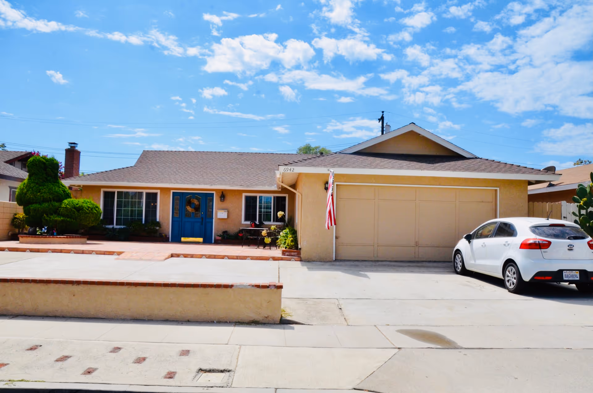 Single-story residential building with a beige exterior, a blue front door, two large windows, a two-car garage, and a white car parked in the driveway. The front yard has a neatly trimmed green shrub and a small patio area with a bench and potted plants. The sky is partly cloudy with blue patches.