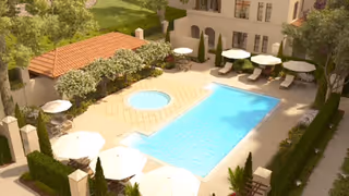 Outdoor swimming pool area with lounge chairs and umbrellas surrounded by greenery and a building in the background.