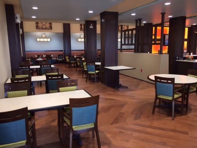 A spacious dining room with multiple tables and chairs arranged neatly. The chairs have wooden frames with blue and green cushions. The room features wooden flooring, dark wooden pillars, and soft lighting from wall-mounted fixtures and ceiling lights. There is a partial divider with illuminated orange panels in the background.