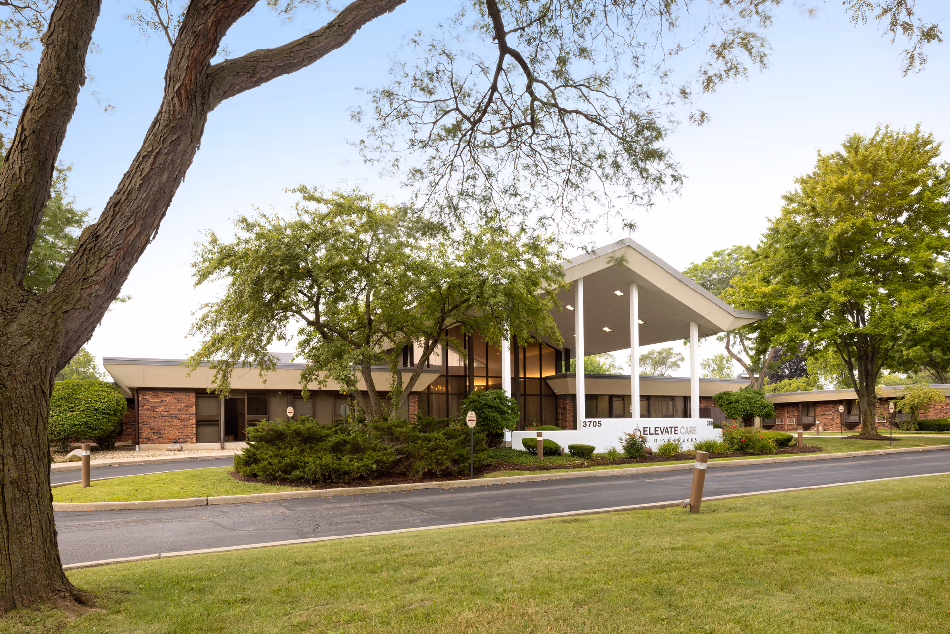 Exterior view of Elevate Care Riverwoods facility showing a modern low-rise building with large windows and a covered entrance supported by white pillars. The building is surrounded by green trees, bushes, and a well-maintained lawn with a driveway in front.
