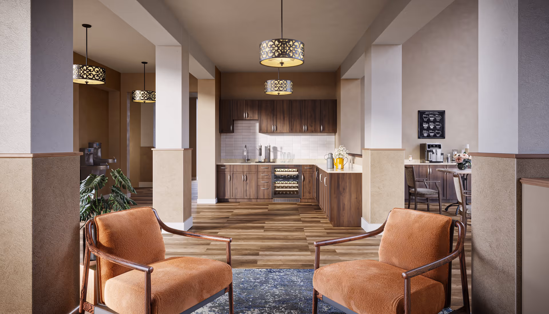Interior view of a senior living facility lounge area with two orange cushioned chairs in the foreground, a kitchen area with wooden cabinets and a wine cooler in the background, pendant lights hanging from the ceiling, and a coffee station with a coffee machine and a framed coffee menu on the wall to the right.