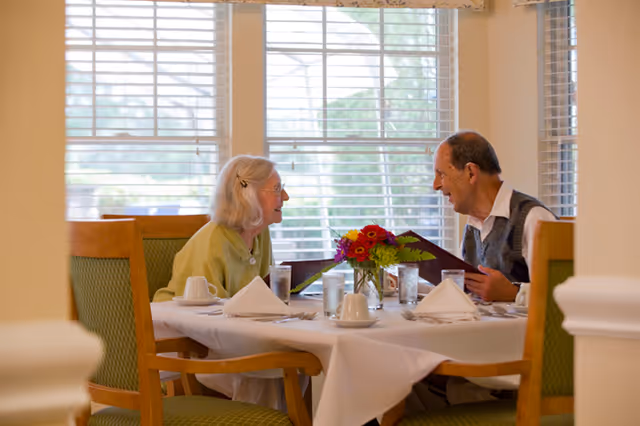 An elderly man and woman sitting at a dining table in a well-lit room with large windows and blinds. The table is set with white tablecloth, napkins, cups, glasses, and a small vase of flowers. They appear to be engaged in conversation and looking at menus.
