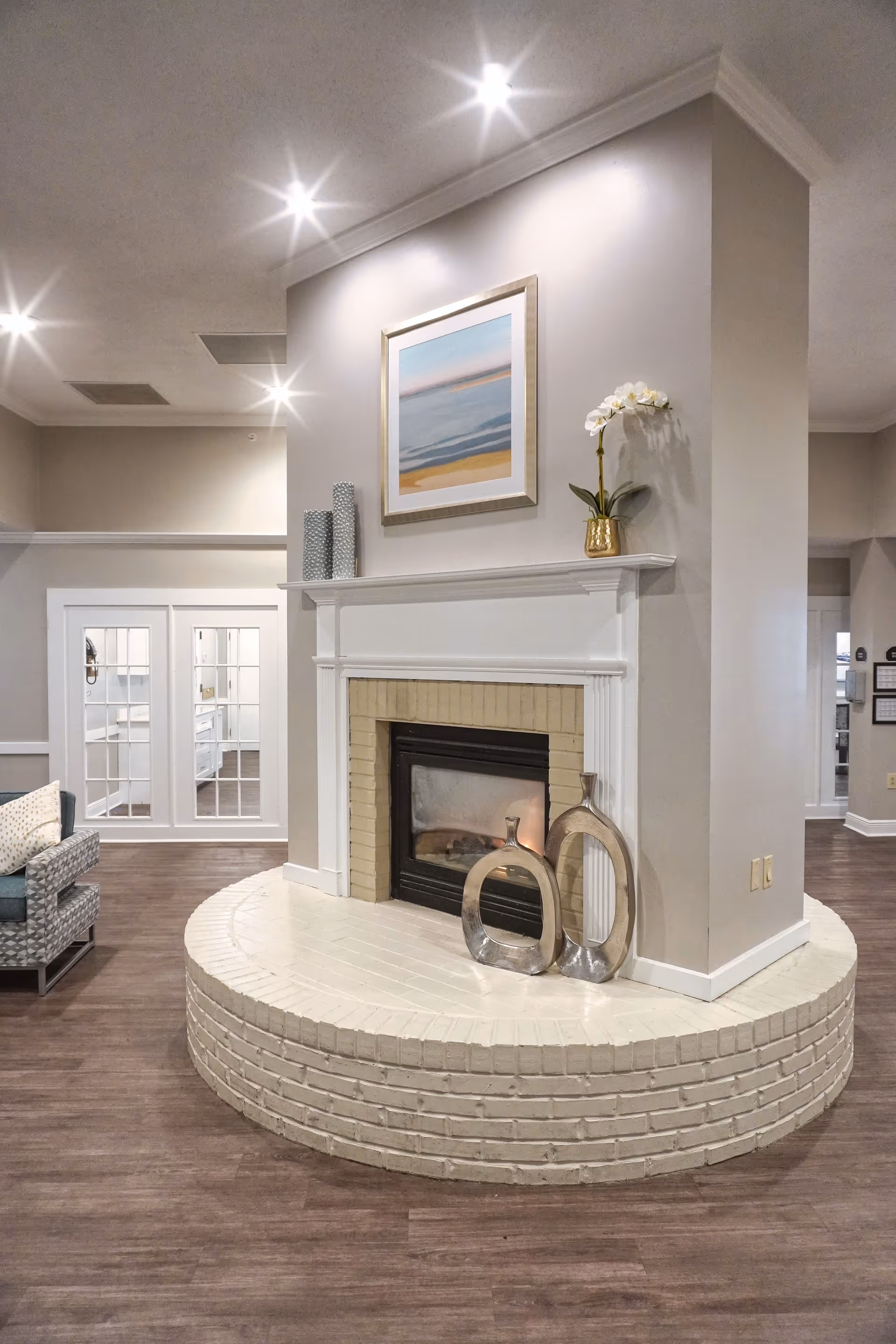 A cozy indoor living area featuring a white brick fireplace with a beige brick interior. On the mantel above the fireplace, there is a framed abstract painting and decorative items including a gold vase with white flowers and two cylindrical vases. In front of the fireplace, there are two modern metallic sculptures. The room has wooden flooring and recessed ceiling lights. To the left, there is a glimpse of a cushioned chair and a set of white French doors leading to another room.