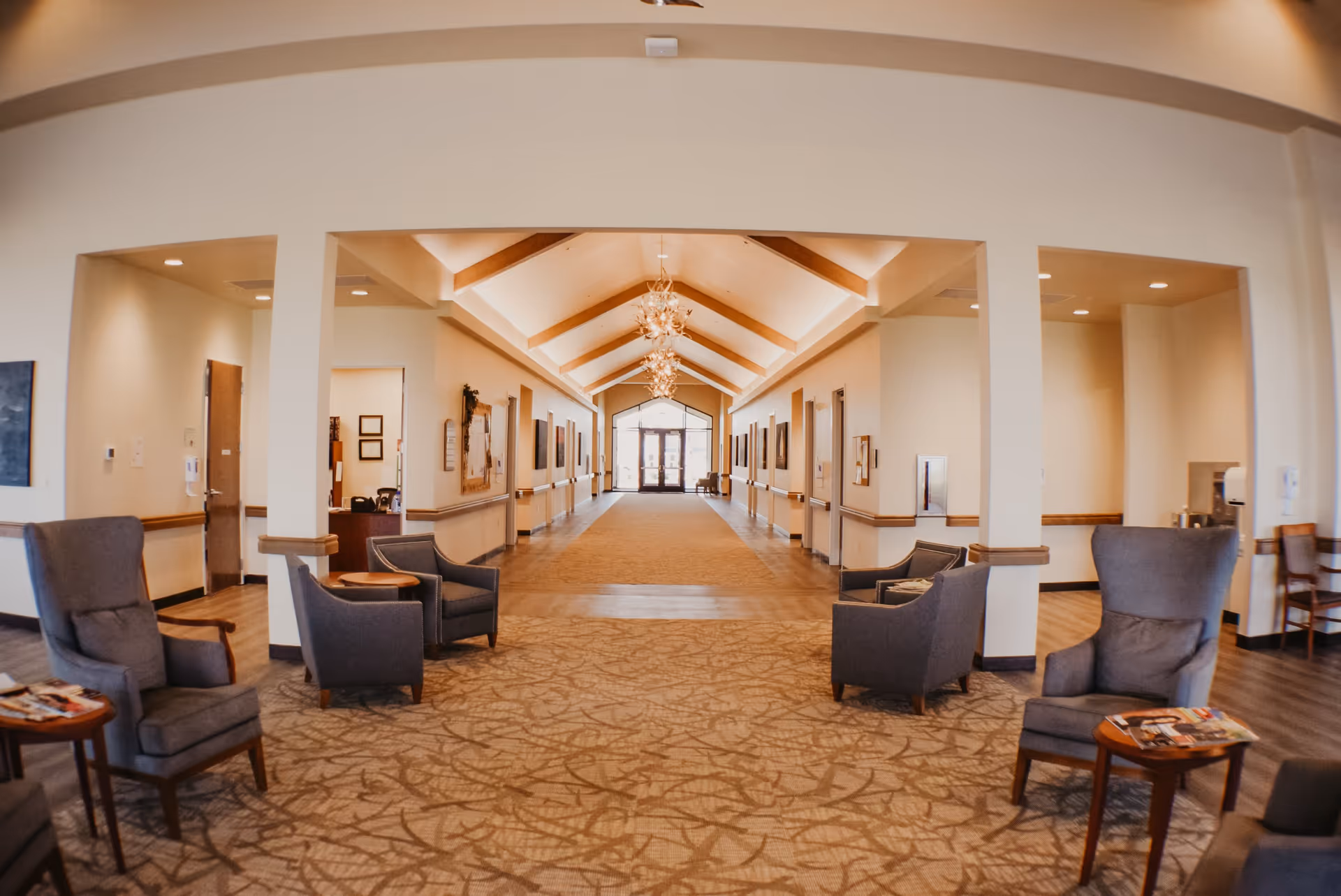 A spacious and well-lit hallway in a senior living facility with a high vaulted ceiling featuring wooden beams and modern chandeliers. The hallway is lined with doors and handrails on both sides, leading to an exit with glass doors at the far end. Comfortable gray armchairs and small wooden tables with magazines are arranged in seating areas near the entrance of the hallway. The walls are painted in a light neutral color, and the floor is carpeted with a patterned design.