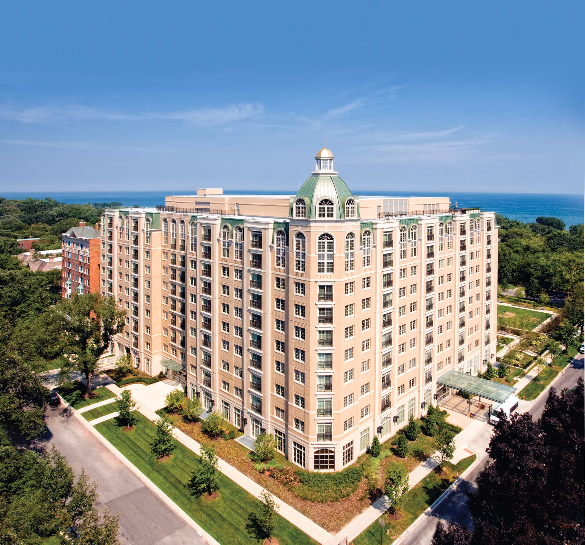 Large multi-story beige residential building with a green-roofed cupola near the waterfront, surrounded by trees and sidewalks.