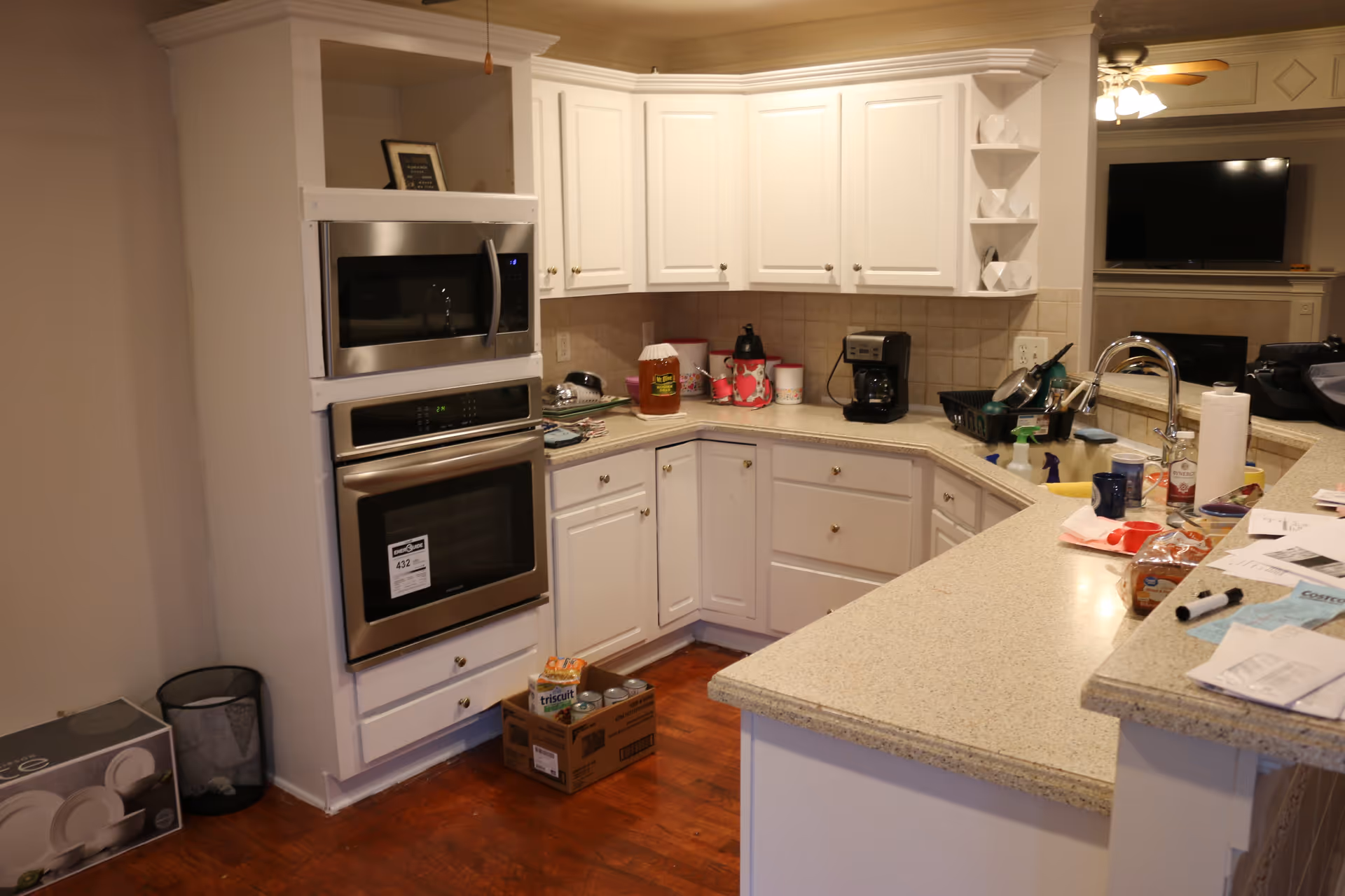 A kitchen area with white cabinets and beige countertops. There is a built-in microwave and oven on the left side. Various kitchen items including a coffee maker, containers, and cleaning supplies are on the countertops. A cardboard box with canned goods and crackers is on the wooden floor. In the background, a living room area with a TV and ceiling fan is partially visible.