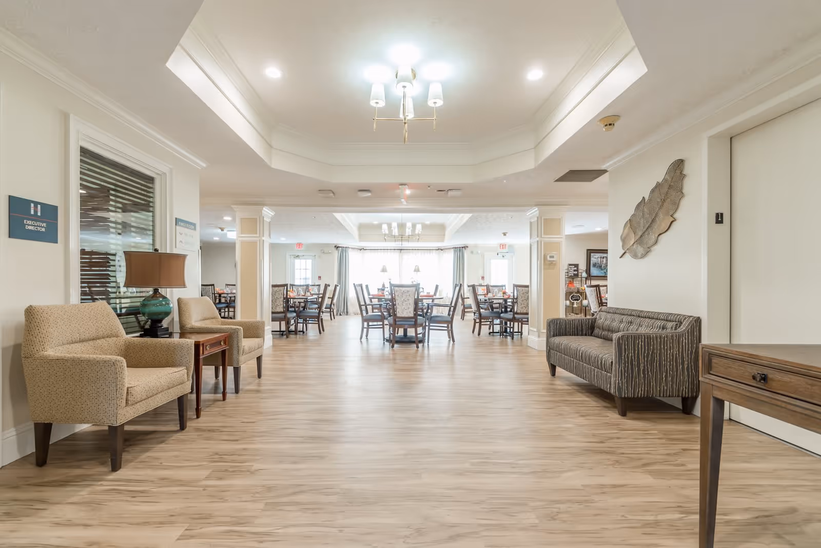 A spacious and well-lit common area in a senior living facility with wooden flooring, beige walls, and ceiling lights. The room features comfortable seating including two beige armchairs with a small table and lamp between them on the left, and a gray patterned sofa on the right. In the background, there are multiple dining tables and chairs arranged neatly near large windows with curtains allowing natural light to fill the space. A decorative leaf wall art is mounted above the sofa, and signs indicating 'Executive Director' and other directions are visible on the walls.