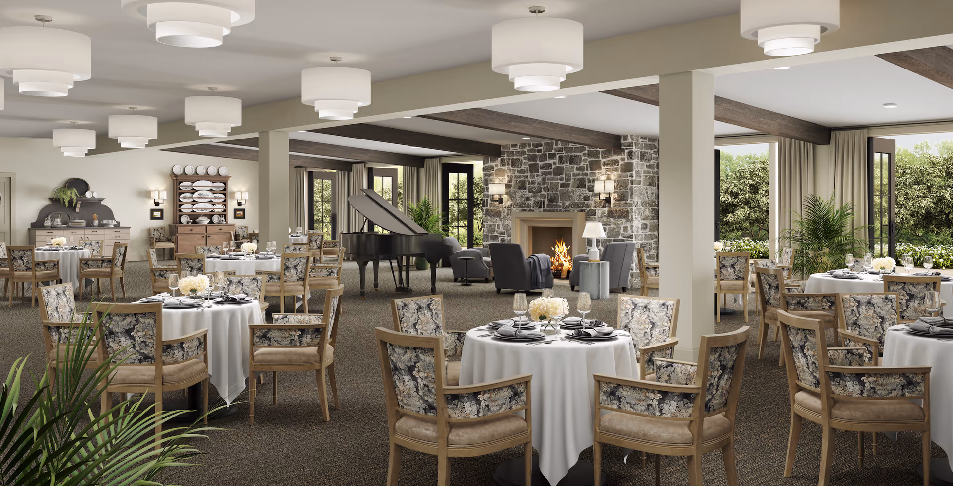 A spacious dining room with round tables covered in white tablecloths, set with plates, glasses, and floral centerpieces. The room features wooden chairs with floral upholstery, a stone fireplace with a fire burning, a grand piano, and large windows with views of greenery outside. The ceiling has modern white light fixtures and wooden beams.