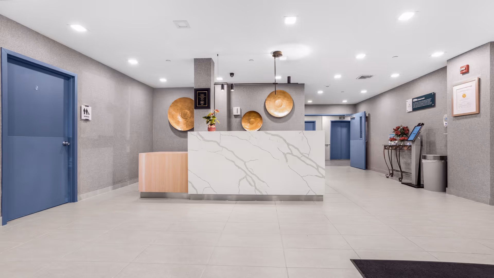 A clean and modern reception area in a healthcare facility with a white marble-patterned front desk, decorative wooden wall hangings, and blue doors. The floor is tiled, and the walls are gray with framed certificates and a small table with flowers and a digital kiosk on the right side.