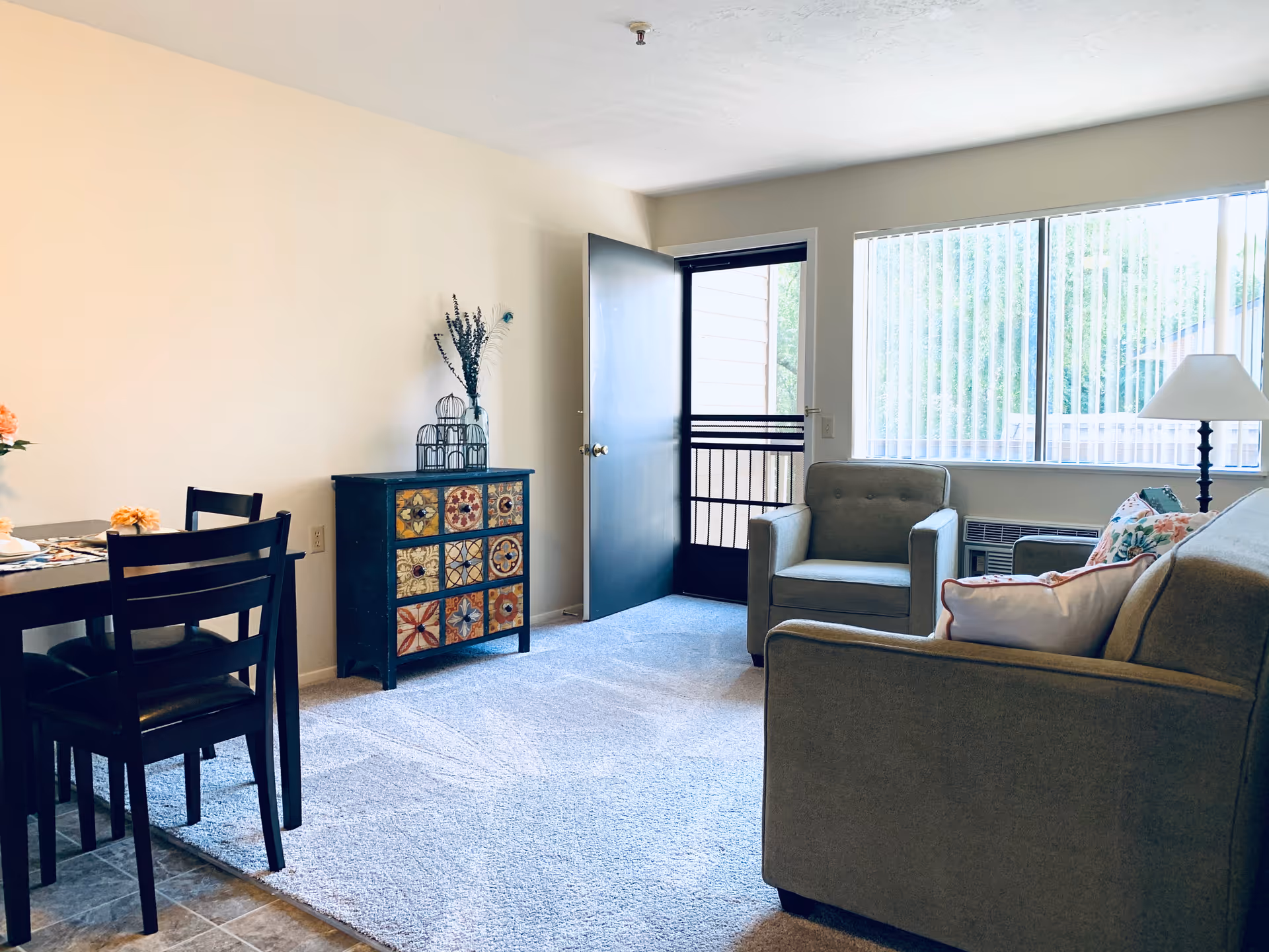 A bright living room with a gray armchair and sofa adorned with floral and white pillows. There is a colorful chest of drawers against a beige wall, a dining table with black chairs on the left, and a floor lamp near a large window with vertical blinds. The door to a balcony or outside area is open, letting in natural light.