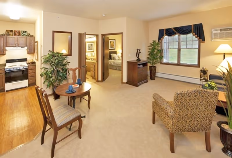 A cozy living area in a senior living facility with a small round wooden table set with two blue cups and saucers, two wooden chairs with striped cushions, a patterned armchair, a wooden cabinet, and a large window with dark blue valance curtains. The room has beige walls and carpet, with a kitchen area featuring wooden cabinets and a stove visible to the left. Two open doorways lead to a bedroom and a bathroom.