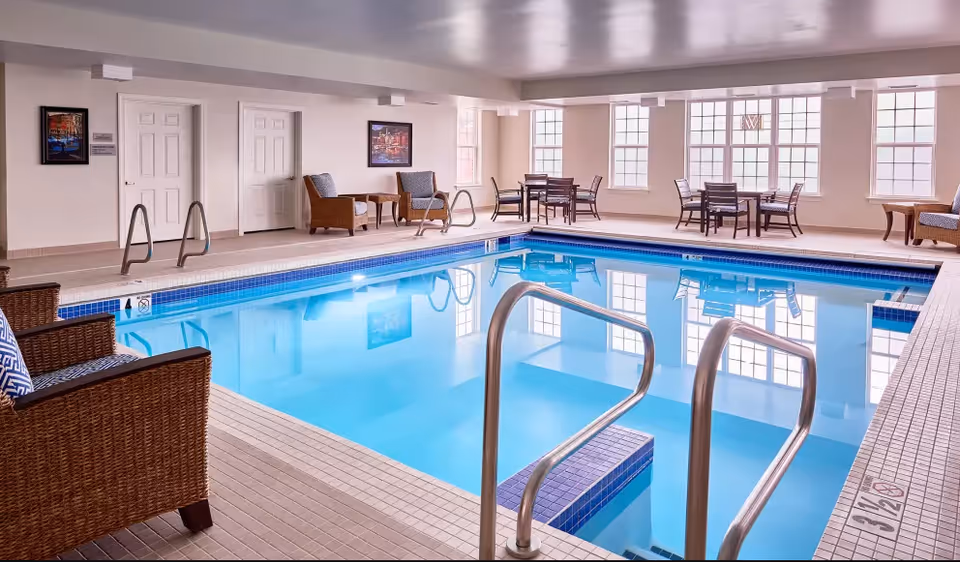 Indoor swimming pool area with clear blue water, surrounded by tiled flooring. Several wicker chairs with cushions and small tables are arranged around the pool. Large windows allow natural light to fill the room, and framed artwork hangs on the walls.