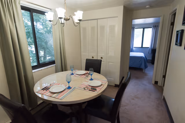 Small dining area with a round table set for four beside a window, with a closet and a bedroom visible down a short hallway.