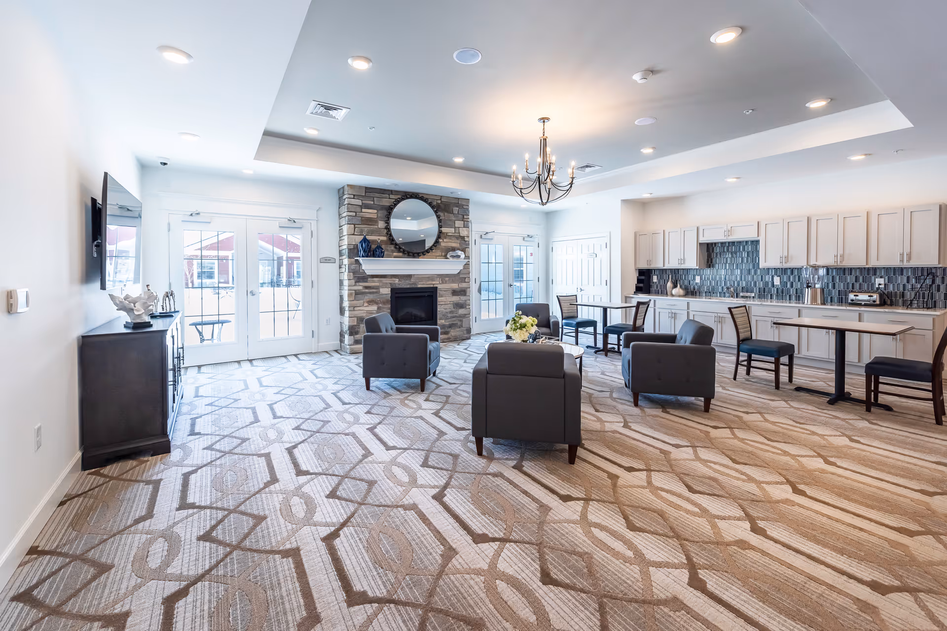A spacious and well-lit common area with patterned carpet flooring, a stone fireplace with a round mirror above it, several gray armchairs arranged around a small table with a flower centerpiece, a chandelier hanging from the ceiling, and a kitchenette with white cabinets and a tiled backsplash along one wall. Double glass doors lead to an outdoor area.