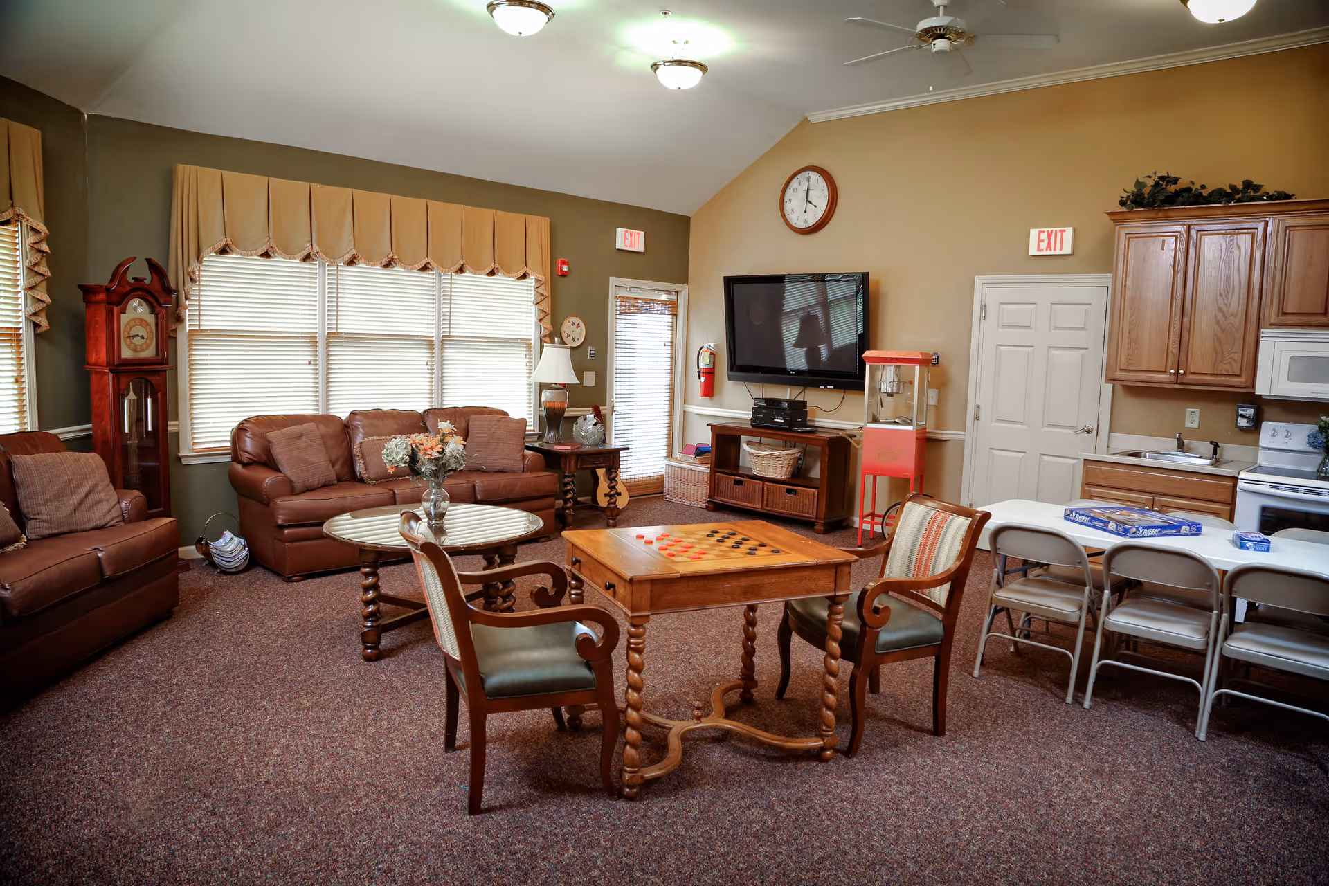 Communal living room with sofas, armchairs, a central game table, TV, and a small kitchenette area.