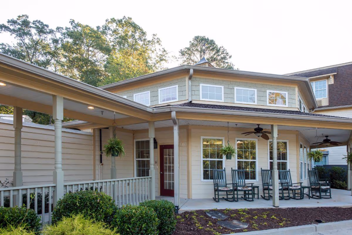 Exterior view of a senior living facility with a covered porch featuring several rocking chairs and hanging plants. The building has multiple windows and is surrounded by greenery and landscaping.