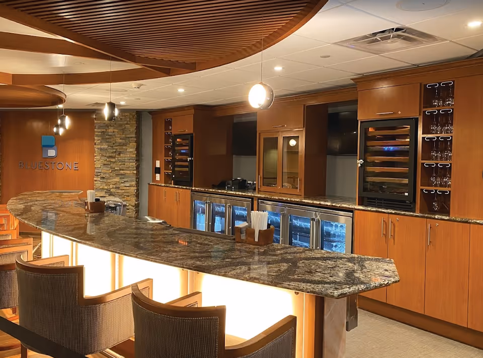 Interior view of a modern bar area with a curved granite countertop, wooden cabinetry, wine coolers, and glassware. The wall features a stone accent and a sign that reads 'BLUESTONE'. Several pendant lights hang from the ceiling, and there are cushioned bar stools along the counter.