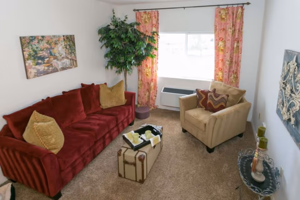 Cozy living room with a red sofa, beige armchair, trunk-style coffee table, potted plant and a window with floral curtains.