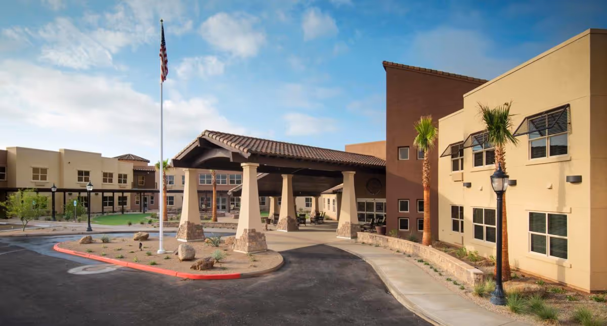 Exterior view of Solana at The Park Assisted Living Community showing the entrance with a covered driveway supported by large columns, a flagpole with an American flag, palm trees, and a clear blue sky.
