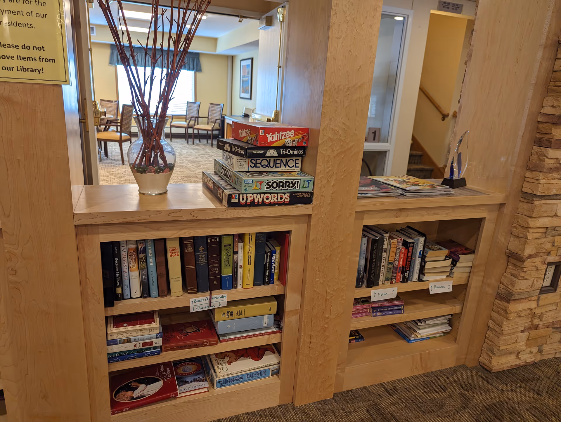 A cozy library area in a senior living facility with wooden bookshelves filled with books and board games like Yahtzee, Sequence, Sorry, and Upwords. A glass vase with decorative branches sits on top of the shelves. In the background, there are chairs arranged near windows with blue valances, and a staircase is visible to the right.