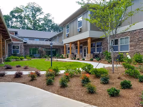 Outdoor courtyard area of a senior living facility with a well-maintained lawn, landscaped garden beds, young trees, and a paved walkway. The building surrounding the courtyard has stone and siding exterior with multiple windows and a covered porch supported by wooden columns.