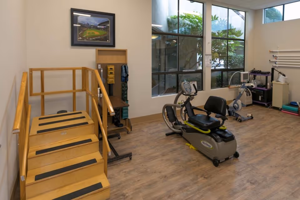 A rehabilitation exercise room with wooden stairs for physical therapy, a recumbent exercise bike, various exercise equipment, and large windows letting in natural light.
