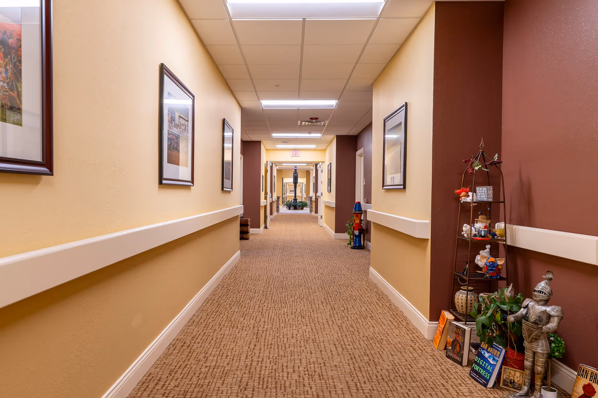 A well-lit hallway in a senior living facility with beige and brown walls, carpeted floor, framed pictures on the walls, and decorative items including a small knight statue, plants, and books on a shelf to the right.