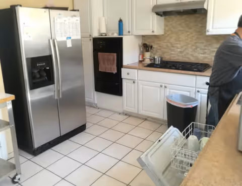 A bright kitchen with a stainless steel side-by-side refrigerator, white cabinets, an open dishwasher, a gas cooktop, and a person standing at the counter.