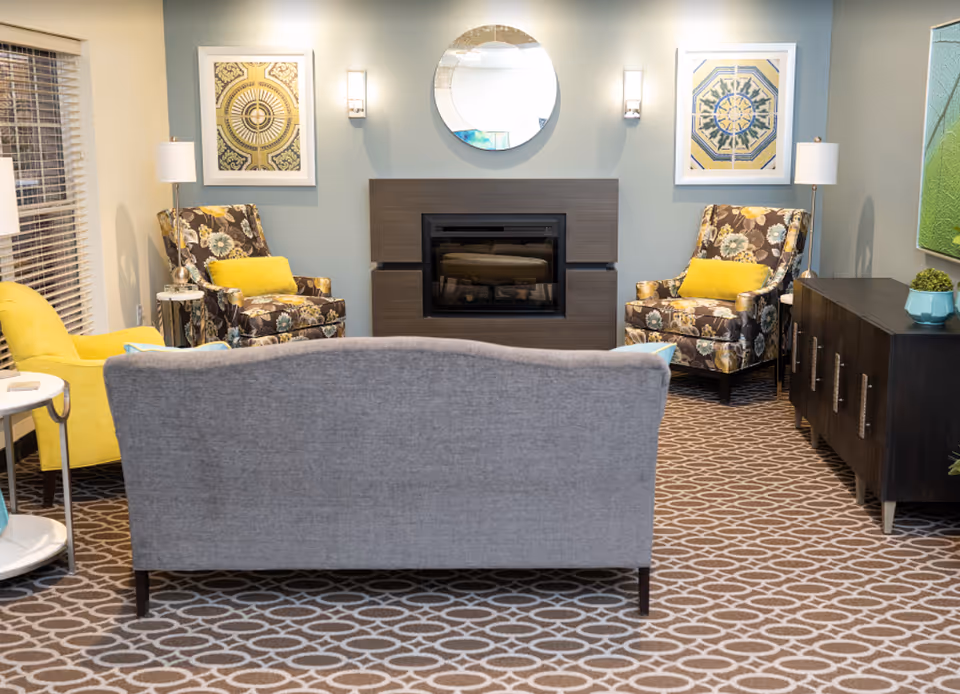 A cozy living room with a gray sofa facing a modern fireplace. There are two floral-patterned armchairs with yellow cushions on either side of the fireplace, and a bright yellow armchair on the left. The room has patterned carpet, a round mirror above the fireplace, framed artwork on the walls, and a dark wooden cabinet on the right side with a small plant on top.