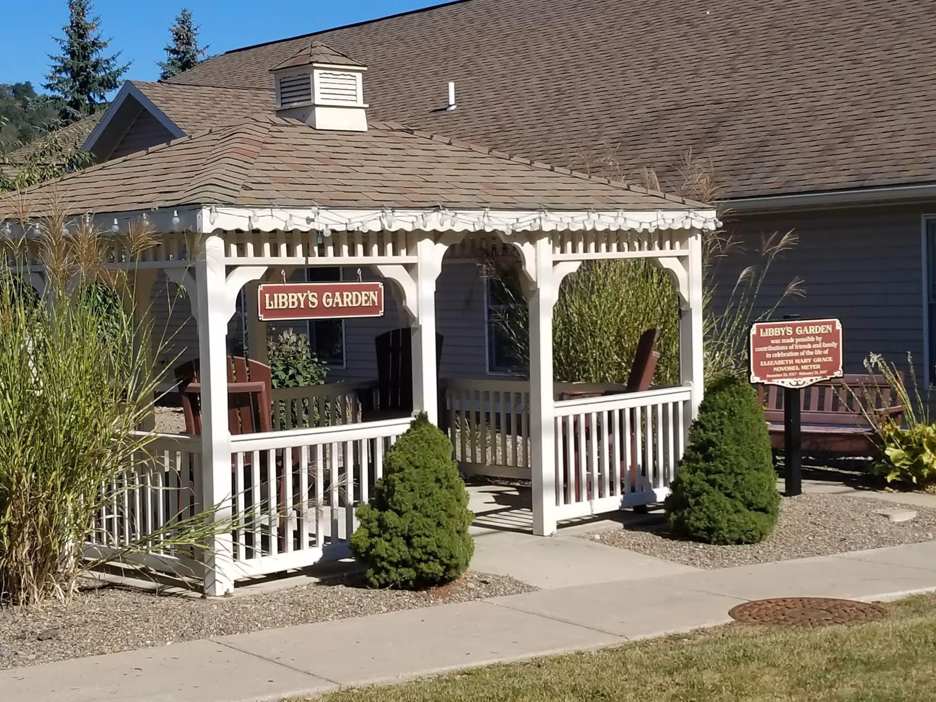 A white wooden gazebo labeled 'Libby's Garden' with a roof and railings, surrounded by small bushes and ornamental grasses. There is a sign next to the gazebo with additional text about Libby's Garden. The gazebo is situated on a concrete path with a building in the background.