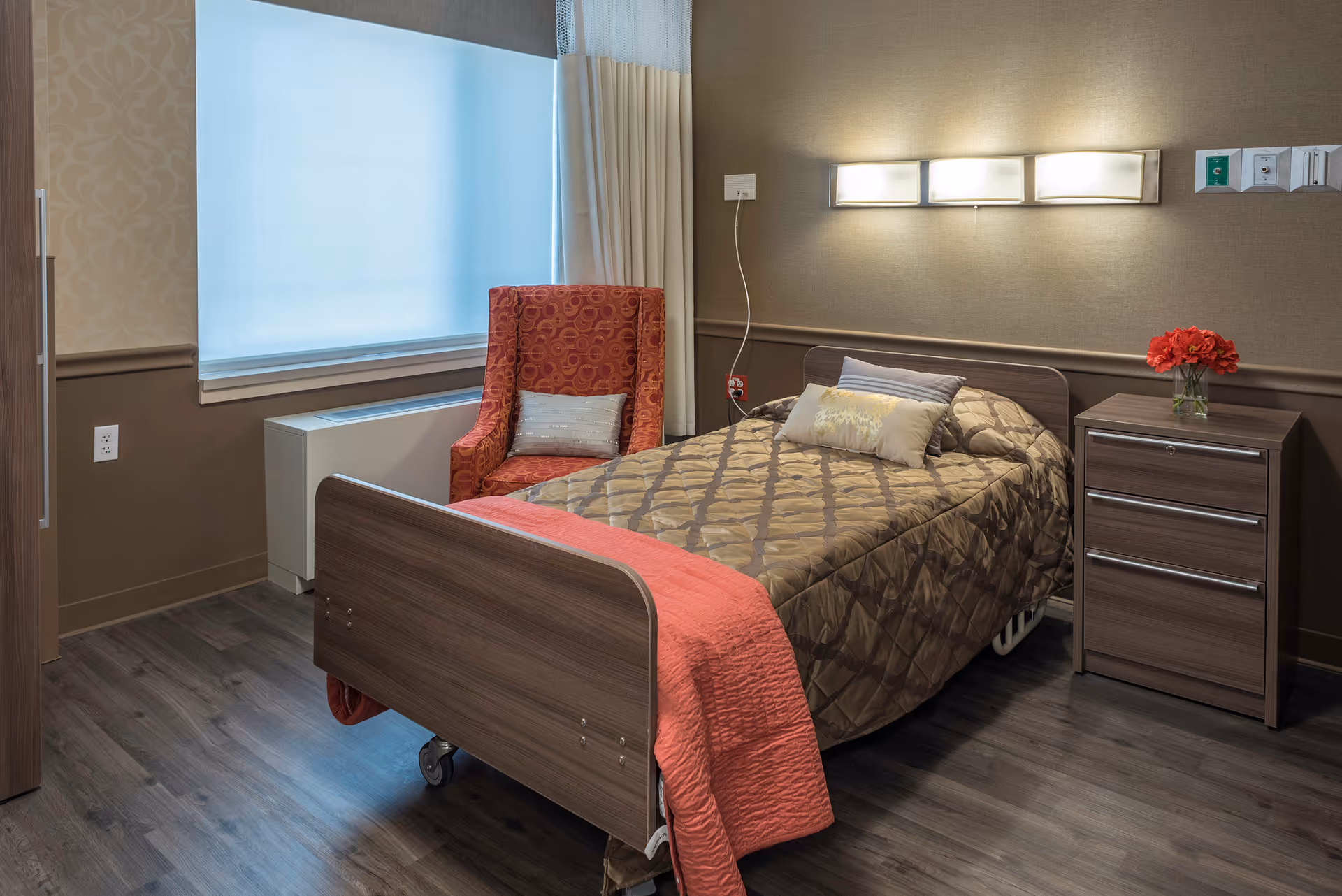 A single bed with a brown patterned bedspread and a coral throw blanket in a room with wooden flooring. Next to the bed is a wooden nightstand with a vase of red flowers. A red patterned armchair with a small pillow is positioned near a large window covered with a sheer white curtain. The wall behind the bed has a modern light fixture and medical equipment outlets.