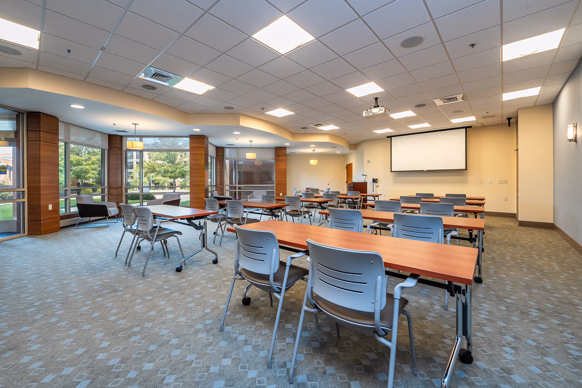 A spacious, well-lit meeting or conference room with multiple rectangular tables and gray chairs arranged in rows facing a large projection screen on the wall. The room has large windows with views of greenery outside, carpeted floors, and a ceiling with recessed lighting and a projector mounted.